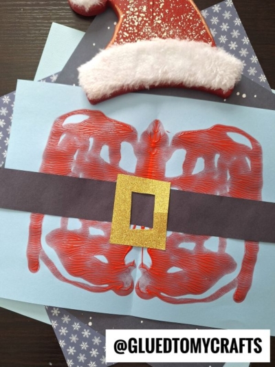 A paint splat Santa in a red suit with a black belt and gold buckle is painted on blue paper, topped with a red felt hat and displayed on snowflake paper. Text at the bottom reads @GLUEDTOMYCRAFTS.