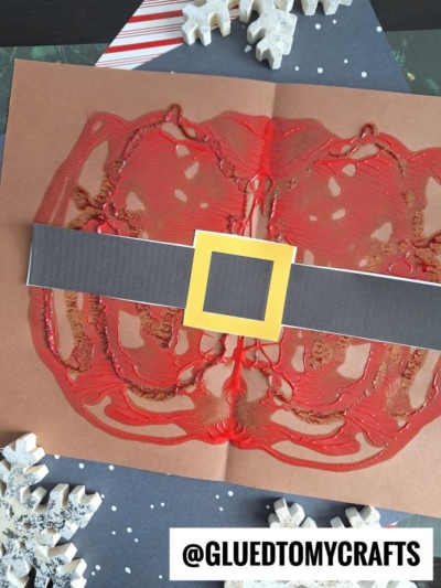 A kid’s craft featuring a paint splat Santa suit with a black belt and yellow buckle, made on brown paper. White snowflake decorations sit nearby, and the handle @GLUEDTOMYCRAFTS is at the bottom.