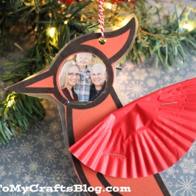 A handmade cardinal photo ornament crafted from paper and a red cupcake liner, featuring a family photo as the bird’s face, hangs on a Christmas tree.