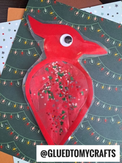 A red cardinal suncatcher craft, shaped like a bird and made from paper and laminate, filled with glitter, and decorated with a googly eye, lies on a green holiday-themed background. @GLUEDTOMYCRAFTS appears at the bottom.