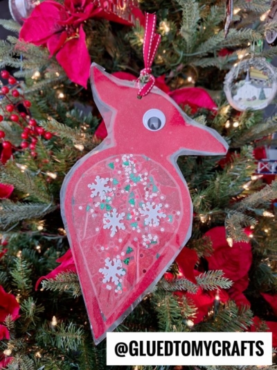 A handmade cardinal suncatcher ornament with a googly eye, decorated with white snowflakes and glitter, hangs on a Christmas tree. The ornament is attached with a red ribbon. Tag: @GLUEDTOMYCRAFTS.