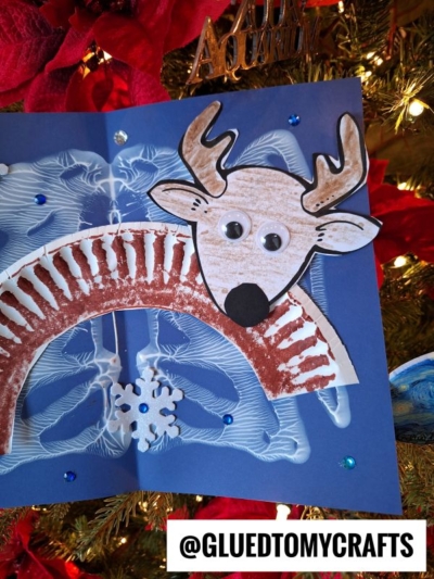 A handmade reindeer craft with googly eyes and brown coloring is displayed against a decorated Christmas tree, featuring ornaments and lights. This Snow Craft Idea For Kids includes a snowflake and blue background. @GLUEDTOMYCRAFTS is labeled at the bottom.