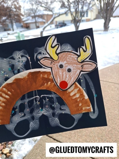 A hand holds a reindeer craft made from a painted paper plate and colored paper, perfect as a Snow Craft Idea For Kids, with snowy houses and trees visible in the background. The text @GLUEDTOMYCRAFTS appears at the bottom.