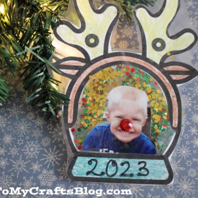 A handmade reindeer photo ornament frame with antlers, googly eyes, and a red glitter nose features a smiling child’s photo labeled 2023. Pine branches and gold stars decorate the background.