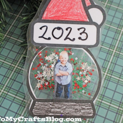 A handmade Santa Snow Globe Christmas ornament with 2023, a Santa hat, and a child's photo inside, hangs on a tree.