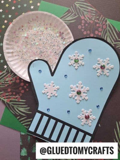 A blue paper mitten decorated with white snowflakes, sequins, and glitter sits on top of festive craft papers and a shimmering paper plate snowball. The handle @GLUEDTOMYCRAFTS is at the bottom.