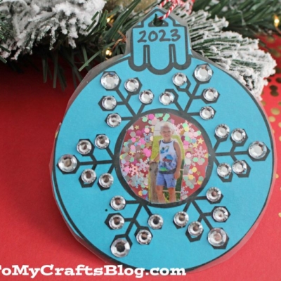 A blue, homemade snowflake photo ornament craft features silver sequins, a central photo of a child, and 2023 written on top. The ornament is displayed on red paper near a decorated tree.