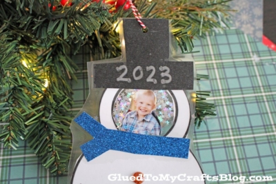 A handmade snowman photo ornament with 2023 on its hat, featuring a smiling child's picture, hangs on a Christmas tree. The snowman has a blue glittery scarf and is set against a green plaid background.