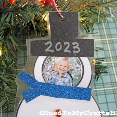 A handmade snowman photo ornament with 2023 on its hat, featuring a smiling child's picture, hangs on a Christmas tree. The snowman has a blue glittery scarf and is set against a green plaid background.
