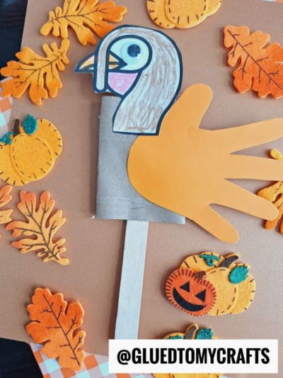 A kids turkey puppet craft made with a paper hand cutout, colored turkey face, googly eye, and a stick—plus inspiration for a classic toilet paper roll turkey—surrounded by felt pumpkins and leaves on a brown background.