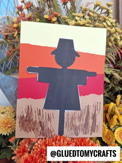 A handmade card featuring a striking scarecrow silhouette cutout, set against a colorful torn-paper sunset background, with brown painted grass and fall flowers artfully surrounding it. Text @GLUEDTOMYCRAFTS is at the bottom.