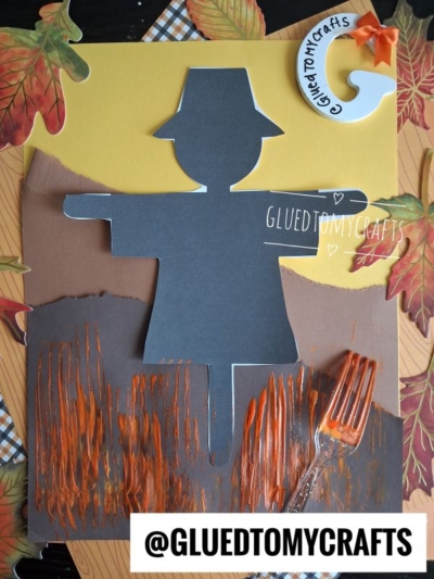 A scarecrow silhouette craft made from cut paper, with brown and orange painted stripes at the bottom, artificial leaves, and a plastic fork on the side. The username @GLUEDTOMYCRAFTS appears twice.