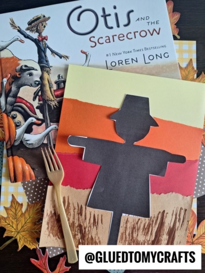 A children’s book titled Otis and the Scarecrow sits beside a scarecrow silhouette paper craft made with torn colored paper strips. A wooden fork and autumn leaves decorate the scene. @GLUEDTOMYCRAFTS is written at the bottom.