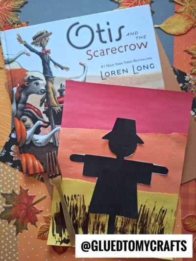 A children’s book titled Otis and the Scarecrow lies next to a handmade Scarecrow Silhouette art project, both placed on autumn-themed paper with artificial leaves. @GLUEDTOMYCRAFTS is written at the bottom.