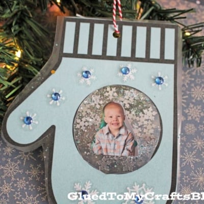 A Mitten Photo Ornament adorned with blue gems and snowflakes hangs near a glowing Christmas tree, framing a smiling child in plaid. The bottom reads “GluedToMyCraftsBlog.com”.