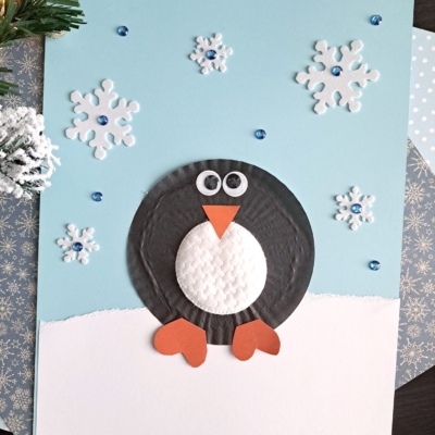 A handmade winter card features a penguin crafted from textured paper in front of a snowy scene, perfect for cupcake liner card crafts. Foam snowflakes and blue gem accents shine on a light blue background. The handle @GLUEDTOMYCRAFTS is shown at the bottom.