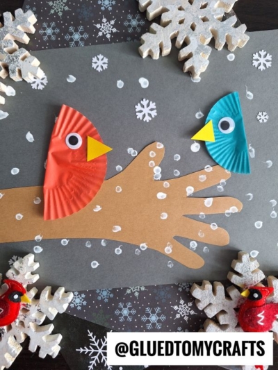 A handmade winter craft featuring two birds made from colored cupcake liners perched on a brown paper hand resembling a q-tip painted winter tree, with white snowflakes and painted snow on a dark background. Text at the bottom reads @GLUEDTOMYCRAFTS.