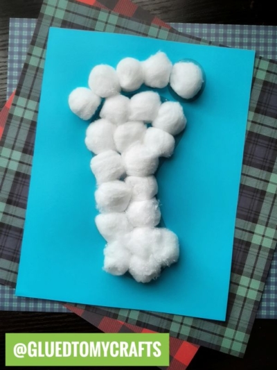 A blue sheet of paper with a letter I made from white cotton balls resembles a playful yeti footprint; the sheet is placed on top of patterned plaid papers. A green label at the bottom reads @GLUEDTOMYCRAFTS.