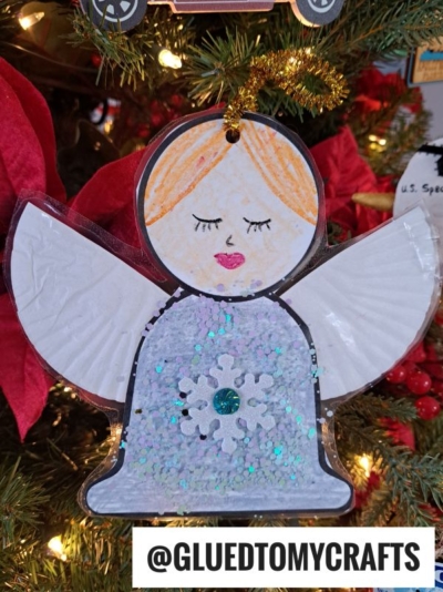 A handmade cupcake liner angel ornament with orange hair, closed eyes, and pink lips hangs on a Christmas tree. The angel has white wings and a glittery blue dress with a snowflake decoration. @GLUEDTOMYCRAFTS is written below.