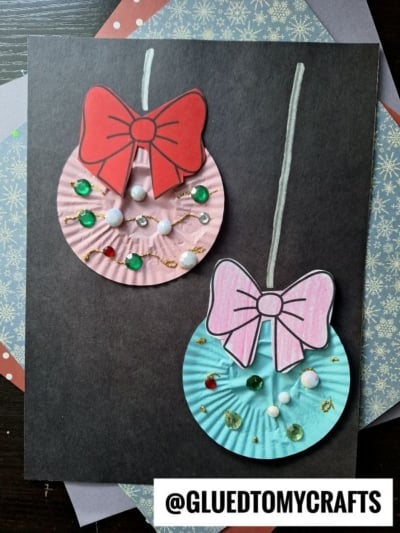 Two colorful Cupcake Liner Ornaments with large bows, sequins, and glitter are displayed on a black background. The pink and blue ornaments are handmade and festive. @GLUEDTOMYCRAFTS is written at the bottom.