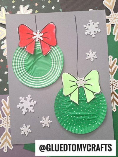 Two green paper plate ornaments with red and green bows are displayed on a gray background, decorated with white snowflake cutouts. Inspired by a Cupcake Liner Ornament, the ornaments hang from drawn black lines. @GLUEDTOMYCRAFTS is written at the bottom.
