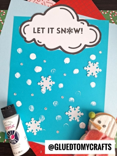 A blue paper features white snowflakes, dots, a cloud reading LET IT SNOW!, a tube of acrylic paint, fingerprint snow accents, a craft snowman, and the handle @GLUEDTOMYCRAFTS at the bottom.