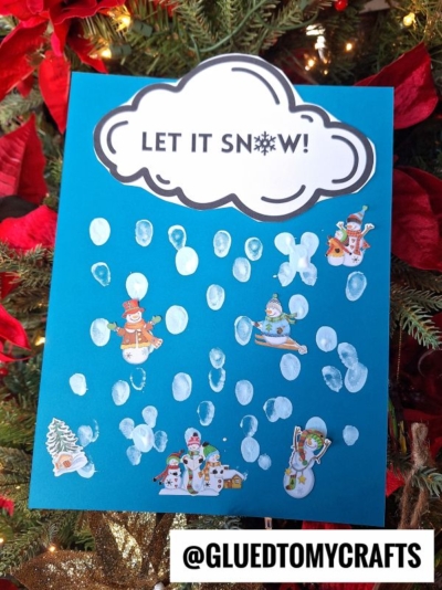 A blue sheet of paper with LET IT SNOW! written in a cloud at the top, decorated with fingerprint snow and winter-themed stickers, displayed in front of a Christmas tree.