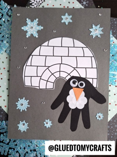 A handmade winter craft features a handprint penguin with googly eyes and a cotton belly, standing by an igloo and surrounded by snowflake stickers on a grey background.