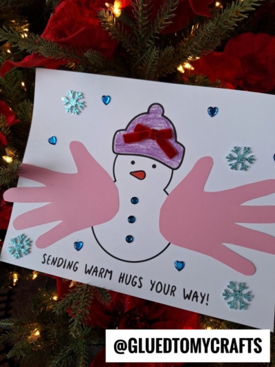 A handmade snowman card on a Christmas tree features pink paper handprints as arms, a purple hat, blue gem buttons, snowflake stickers, and the words SENDING WARM HUGS YOUR WAY! at the bottom—a perfect warm hugs keepsake.