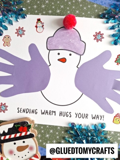 A handmade snowman card with purple paper handprints as arms, a pink pom-pom on its purple hat, and the text Sending warm hugs your way!—a perfect keepsake surrounded by festive stickers.