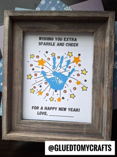Framed artwork featuring a blue handprint surrounded by yellow and orange stars, with the text “Wishing you extra sparkle and cheer for a Happy New Year! Love, _______.” A perfect sparkle and cheer keepsake. Handle @gluedtomycrafts is at the bottom.