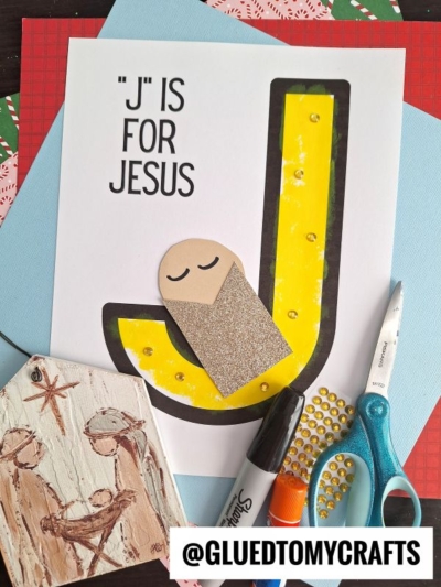 A craft project features a large yellow letter J and a simple baby Jesus figure, surrounded by art supplies, a marker, scissors, and a nativity ornament. The text celebrates "'J' Is For Jesus.