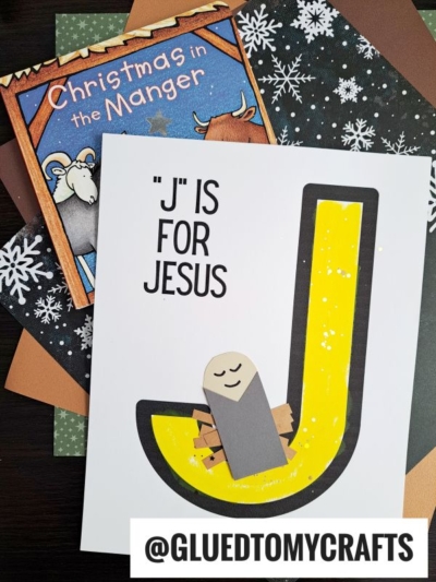 A craft project titled "J" Is For Jesus features a large yellow letter J with a paper baby Jesus in a manger on snowflake-patterned paper. The children's book Christmas in the Manger is also included in the display.