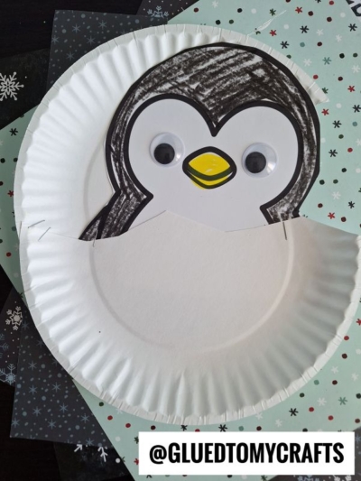 A paper plate hatching penguin craft featuring a cute penguin with googly eyes and a yellow beak, peeking out from behind a cut paper plate, displayed on patterned paper. @GLUEDTOMYCRAFTS is written at the bottom.