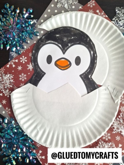 A paper plate hatching penguin peeks out from its egg, displayed on snowflake-patterned paper with blue and silver snowflake decorations. @GLUEDTOMYCRAFTS is written at the bottom for a creative winter touch.