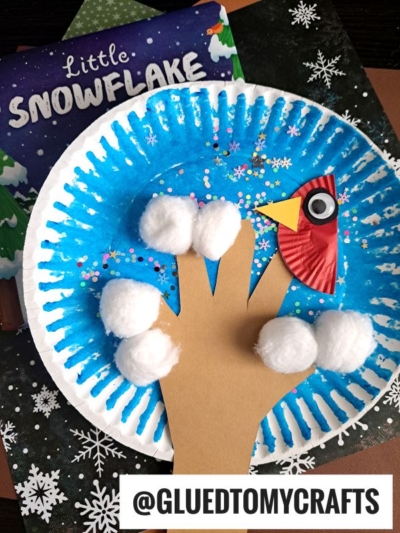 A blue paper plate displays a whimsical winter scene, featuring a red cupcake liner bird perched on a paper hand cutout that serves as a paper plate winter tree. Soft white cotton balls create the look of falling snow, and glitter adds extra sparkle. In the background, a children’s book titled Little Snowflake completes this festive setting.
