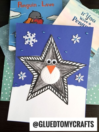 A handmade paper star penguin card features a star-shaped black and white body, googly eyes, and an orange beak on a snowy background, surrounded by snowflake stickers and penguin-themed books in the background.