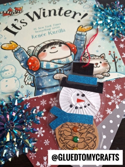 A children’s winter book titled “It’s Winter!” is shown with a handmade pinecone snowman ornament made of paper, glitter, and a red string, along with blue snowflake decorations on a snowflake-patterned background.