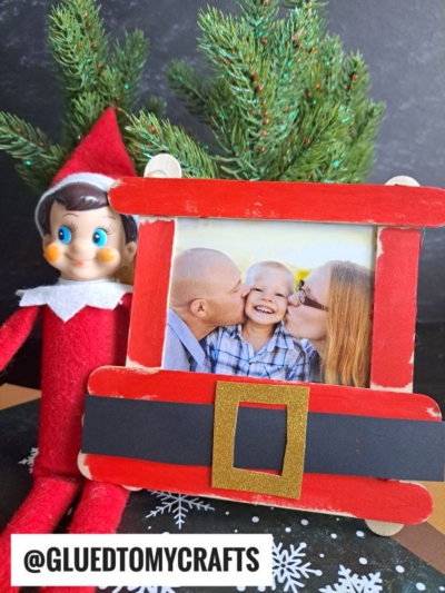 A popsicle stick Christmas frame, decorated like Santa’s suit, holds a family photo. An Elf on the Shelf doll sits beside it, with a small artificial Christmas tree in the background. @GLUETOMYCRAFTS.
