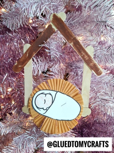 A handmade nativity ornament made of craft sticks and a cupcake liner, showing baby Jesus in a manger, hangs on a pink Christmas tree. This charming piece doubles as a unique nativity photo frame, beautifully capturing the spirit of the season. The text @GLUEDTOMYCRAFTS appears at the bottom.