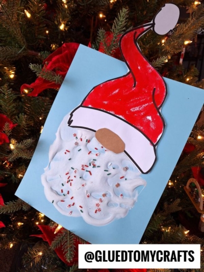 A handmade puffy paint Santa craft with a red hat and a puffy white beard decorated with sprinkles, displayed on blue paper in front of a lit Christmas tree. @GLUEDTOMYCRAFTS is printed at the bottom.