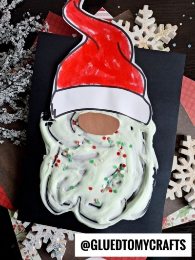 A handmade puffy paint Santa craft with a painted red hat, a white fluffy beard decorated with small beads, and a cut-out face, displayed on a black background with snowflake decorations. @GLUEDTOMYCRAFTS is written at the bottom.