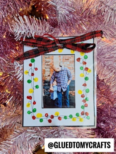 A handmade photo ornament hangs on a pink Christmas tree, featuring a picture of an adult and child. Decorated with Q-Tip Painted Christmas Lights, colorful dots, and a plaid ribbon, it’s finished with @GLUEDTOMYCRAFTS at the bottom.