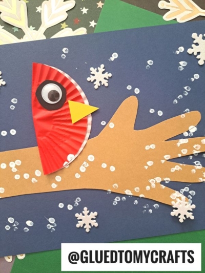 A handmade craft shows a brown handprint cutout on dark blue paper, decorated as a bird with a red cupcake liner wing, googly eye, yellow beak, and snowy accents like q-tip painted winter tree branches and white dots. @GLUEDTOMYCRAFTS is at the bottom.