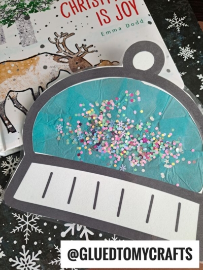 A homemade paper snow globe craft with colorful confetti sits on a snowflake-patterned surface next to a book featuring a reindeer and a charming winter hat suncatcher. Text reads @GLUEDTOMYCRAFTS.