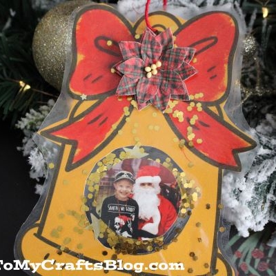 A handmade Christmas Bell Photo Ornament with a large red bow, gold glitter, and a child’s photo with Santa inside. It hangs amid festive greenery and gold decorations.