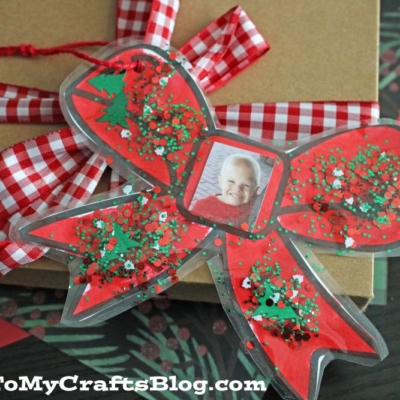 A handmade Christmas Bow Ornament with red, green, and white glitter features a child’s photo at the center and is tied to a gift wrapped in brown paper with a red gingham ribbon.