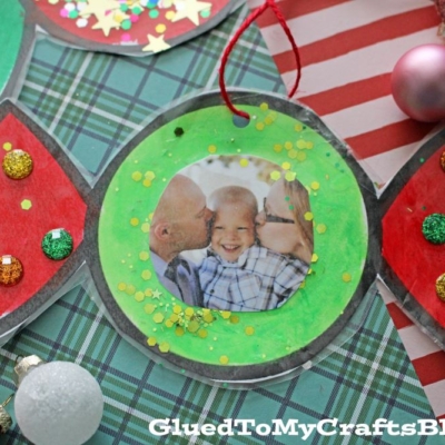 This handmade Candy Photo Ornament is shaped like a wrapped treat, adorned with glitter and gems, and showcases a smiling child's photo at the center. It rests on festive plaid and striped paper for a cheerful holiday touch.