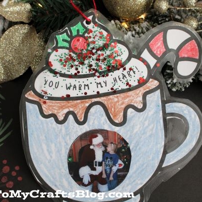 A handmade christmas mug photo ornament shaped like hot chocolate with a candy cane and whipped cream. A photo and “YOU WARM MY HEART” are on the mug, with pine branches and gold ornaments in the background.