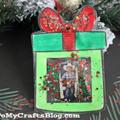 A handmade Christmas Present Photo Ornament shaped like a green gift with a red bow, filled with glitter and featuring a child's photo in the center.